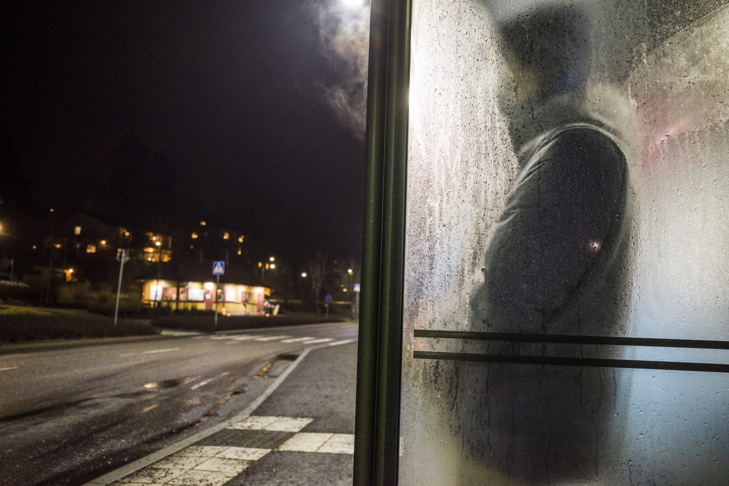 En man står och väntar på bussen i en busskur en mörk och regnig kväll.